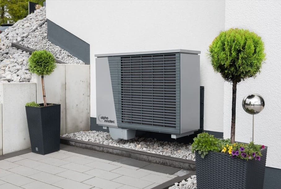 Une pompe à chaleur extérieure de couleur grise est installée à côté d'un bâtiment moderne, entourée de plantes en pot, de gravier et d'un sol carrelé.