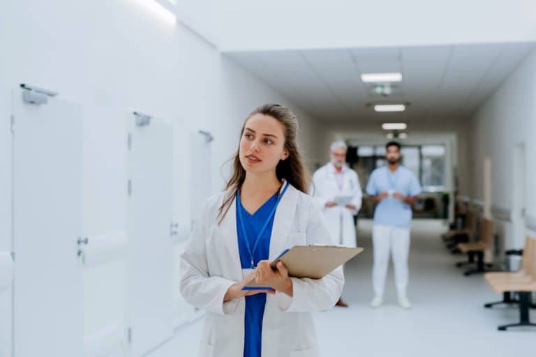 Une femme médecin en blouse de laboratoire tient un presse papiers dans un couloir d'hôpital, avec deux médecins masculins debout en arrière plan.