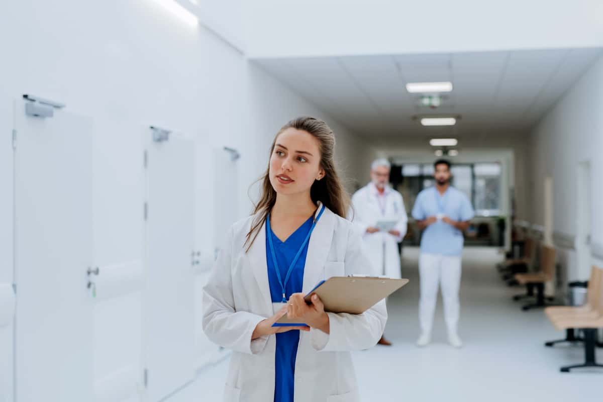 Une femme médecin en blouse de laboratoire tient un presse papiers dans un couloir d'hôpital, avec deux médecins masculins debout en arrière plan.