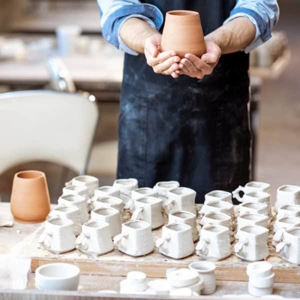 Une personne portant un tablier tient un pot en argile fini au dessus d'une table remplie de nombreuses pièces de poterie inachevées dans un atelier de céramique.