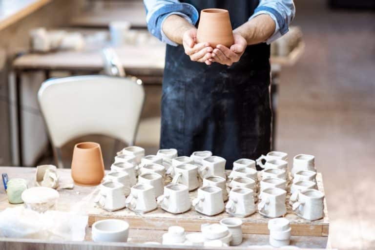 Une personne portant un tablier tient un pot en argile fini au dessus d'une table remplie de nombreuses pièces de poterie inachevées dans un atelier de céramique.