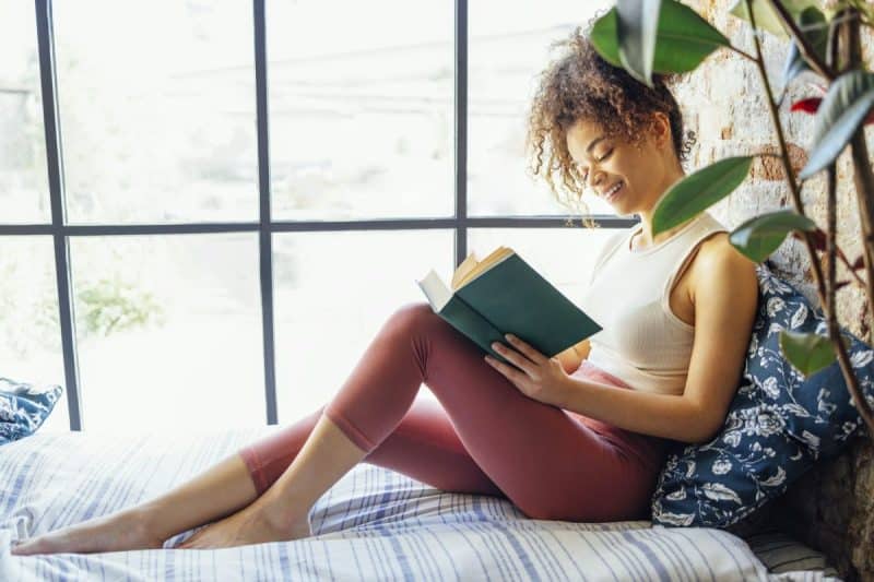 Pretty african american girl reading book sitting on windowsill