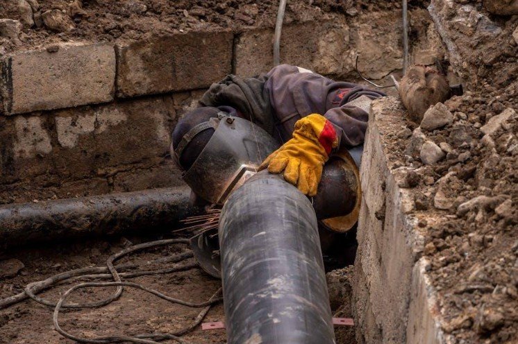 Un ouvrier en tenue de protection soude un gros tuyau métallique dans une tranchée entourée de terre et de gravats de béton.