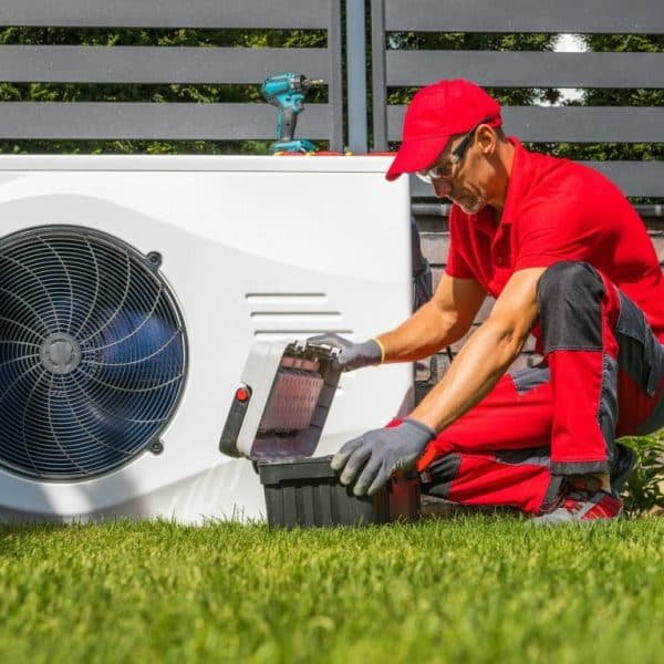 Un technicien portant un uniforme rouge et un chapeau travaille sur une unité CVC extérieure, ouvrant une boîte à outils sur l'herbe.