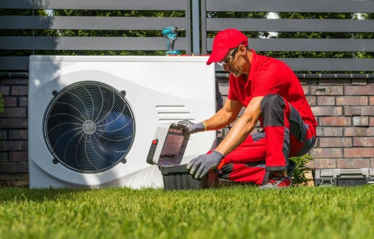 Un technicien portant un uniforme rouge et un chapeau travaille sur une unité CVC extérieure, ouvrant une boîte à outils sur l'herbe.