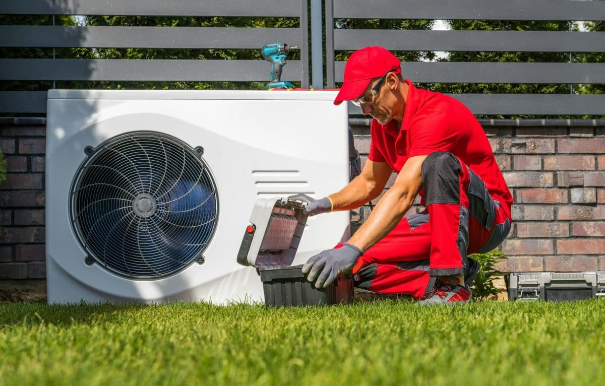 Un technicien portant un uniforme rouge et un chapeau travaille sur une unité CVC extérieure, ouvrant une boîte à outils sur l'herbe.