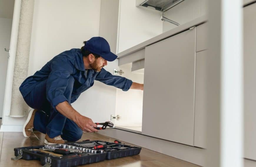 Un plombier en uniforme bleu et casquette examine les tuyaux sous un évier de cuisine, une clé à molette à la main. Une boîte à outils avec des outils est ouverte sur le sol à proximité.