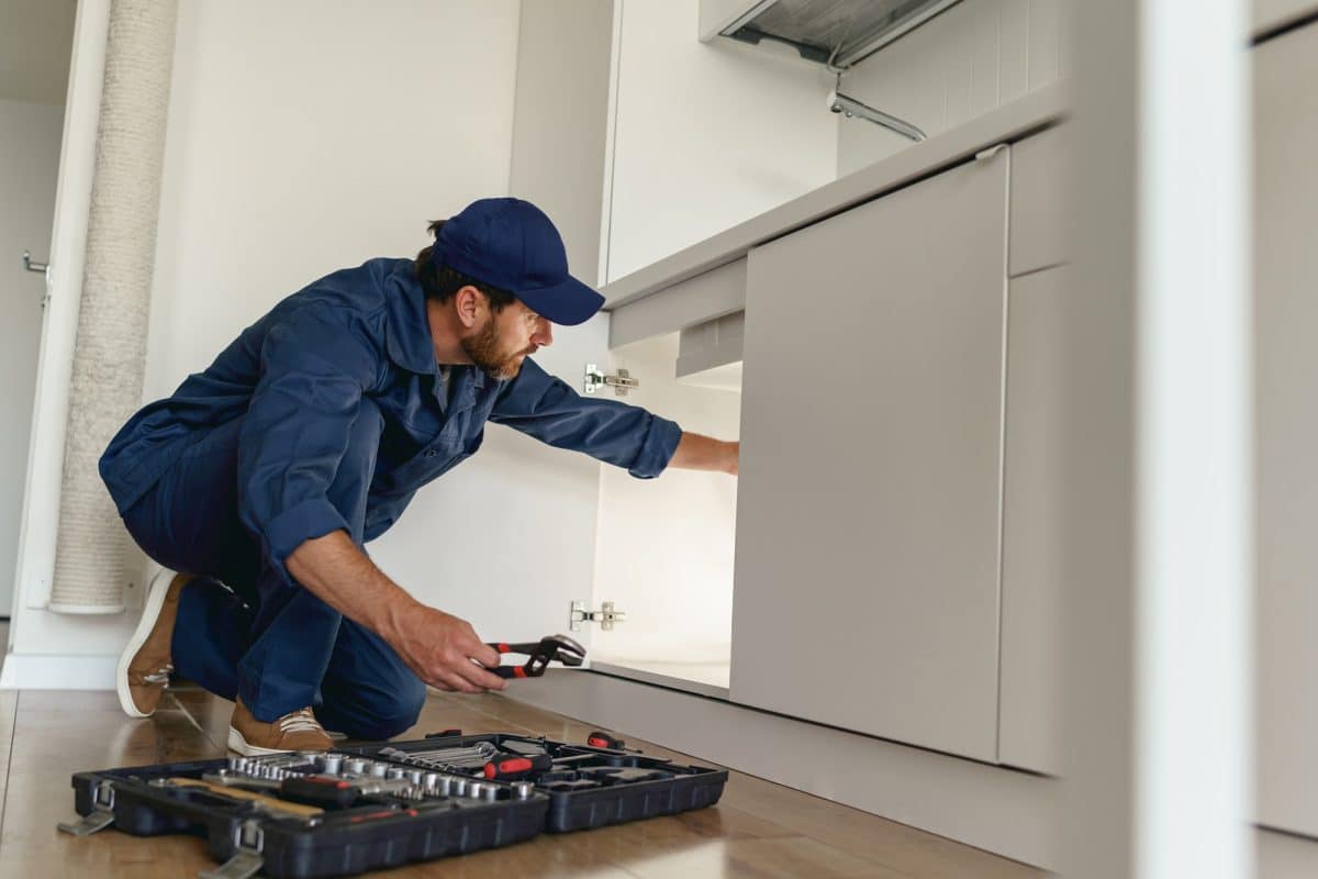Un plombier en uniforme bleu et casquette examine les tuyaux sous un évier de cuisine, une clé à molette à la main. Une boîte à outils avec des outils est ouverte sur le sol à proximité.