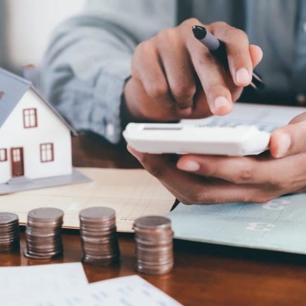 Personne utilisant une calculatrice avec une maison modèle, des pièces empilées et des documents sur une table, suggérant une planification financière ou une budgétisation.