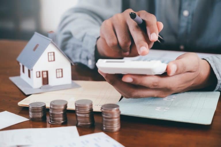 Personne utilisant une calculatrice avec une maison modèle, des pièces empilées et des documents sur une table, suggérant une planification financière ou une budgétisation.