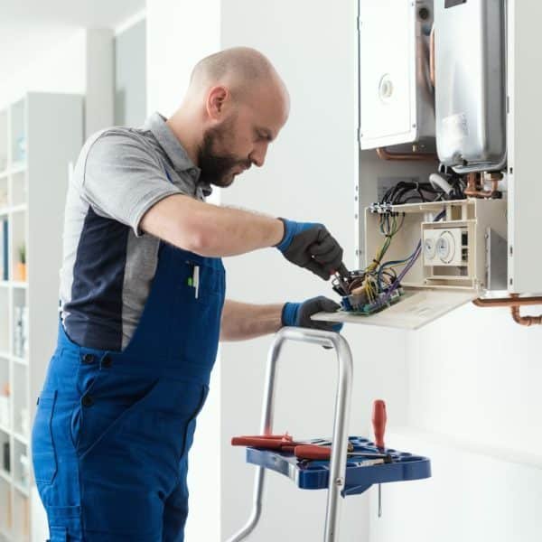 Un technicien en bleu de travail répare une chaudière murale ouverte à l'aide d'outils, debout sur un escabeau dans une pièce lumineuse.