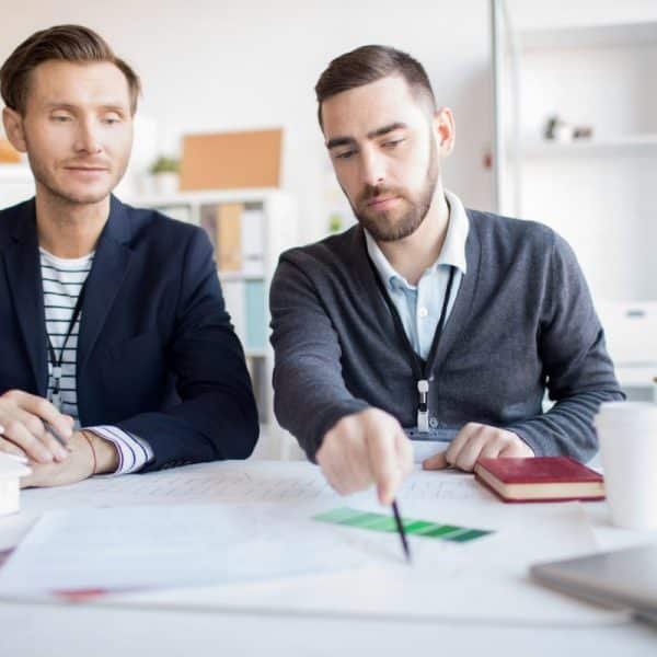 Deux hommes en tenue professionnelle discutent de projets assis à un bureau avec des papiers, un ordinateur portable et une plante en pot. L'un désigne un échantillon de couleur tandis que l'autre observe.