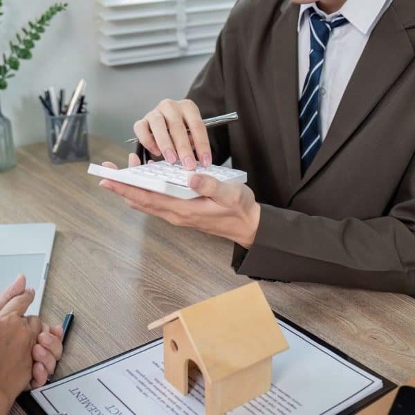 Une personne en costume tient une calculatrice, assise à un bureau avec une petite maquette de maison en bois, un document et un ordinateur portable.