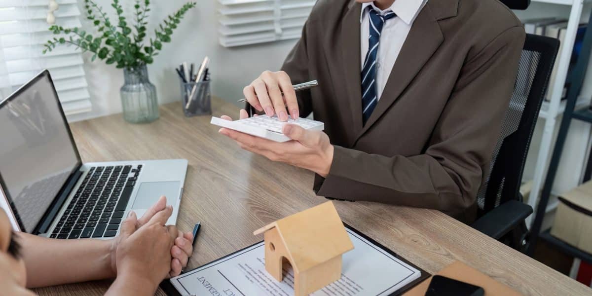 Une personne en costume tient une calculatrice, assise à un bureau avec une petite maquette de maison en bois, un document et un ordinateur portable.