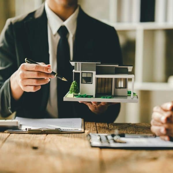 Une personne en costume tient une petite maquette architecturale d'une maison moderne tout en discutant avec une autre personne à une table en bois. Des documents et une calculatrice sont également présents sur la table.