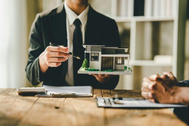 Une personne en costume tient une petite maquette architecturale d'une maison moderne tout en discutant avec une autre personne à une table en bois. Des documents et une calculatrice sont également présents sur la table.