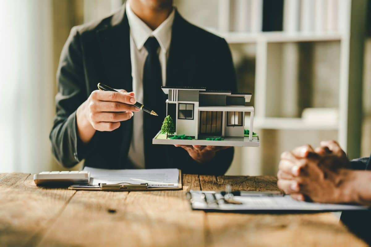 Une personne en costume tient une petite maquette architecturale d'une maison moderne tout en discutant avec une autre personne à une table en bois. Des documents et une calculatrice sont également présents sur la table.