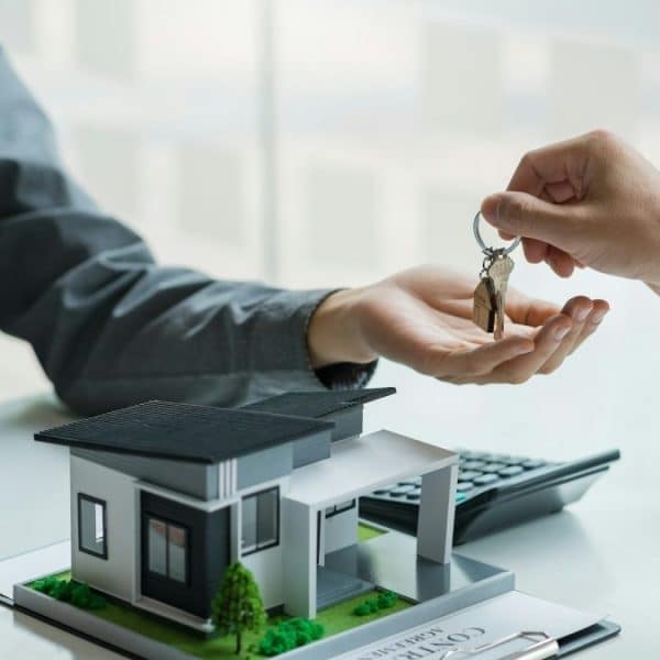 Une main donne les clés de la maison à une autre main au dessus d'une table avec une maquette de maison, un contrat, une calculatrice et un ordinateur portable.