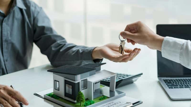 Une main donne les clés de la maison à une autre main au dessus d'une table avec une maquette de maison, un contrat, une calculatrice et un ordinateur portable.