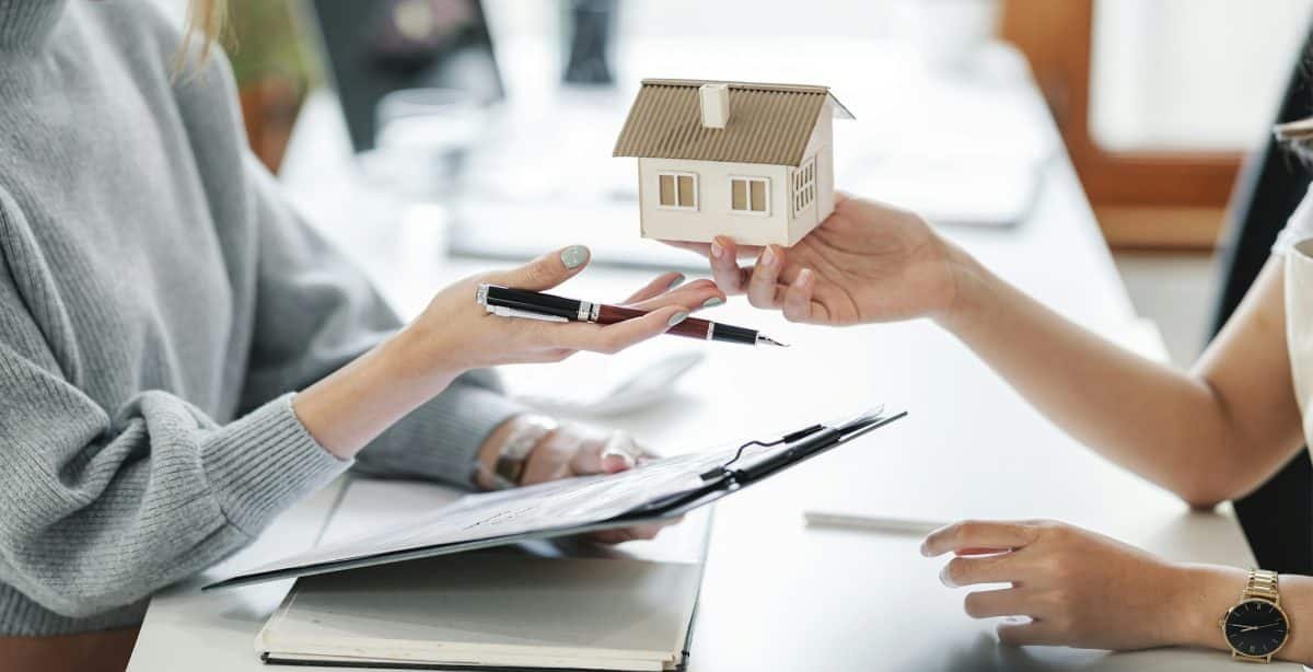 Deux personnes échangent une maquette de maison sur un bureau, l'une d'elles tenant un presse papiers et un stylo.