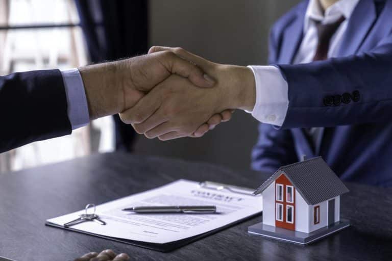 Deux personnes vêtues d'une tenue professionnelle se serrent la main autour d'une table avec un contrat, un stylo et une petite maquette de maison.