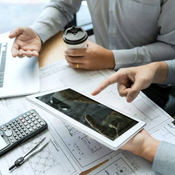 Deux personnes examinent des documents avec une tablette et un ordinateur portable sur une table. Une calculatrice et un stylo sont à proximité, tandis qu'une personne tient une tasse de café.