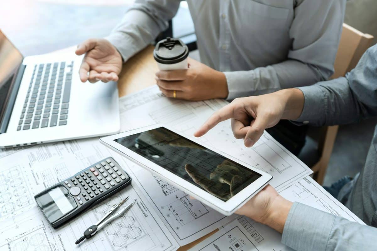 Deux personnes examinent des documents avec une tablette et un ordinateur portable sur une table. Une calculatrice et un stylo sont à proximité, tandis qu'une personne tient une tasse de café.