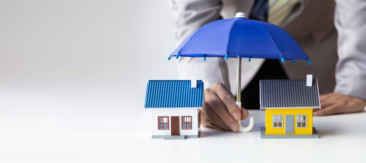 Une personne tient un parapluie bleu au dessus de deux maisons modèles, symbolisant la protection ou l'assurance des maisons.