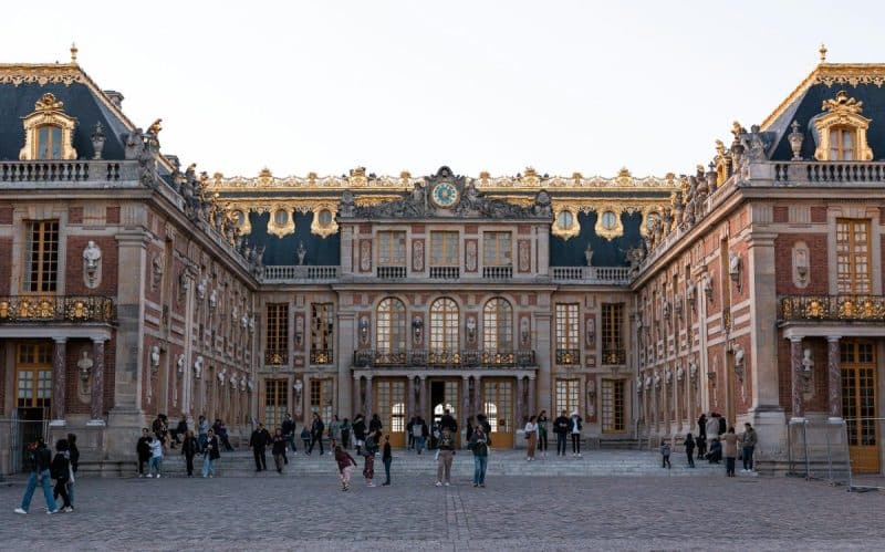 Des gens marchant devant l'extérieur orné du château de Versailles sous un ciel clair.