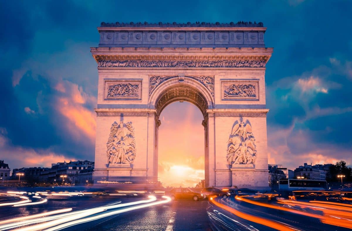 L'Arc de Triomphe au coucher du soleil avec des traînées de phares de voiture, sur fond de ciel dramatique avec des nuages.