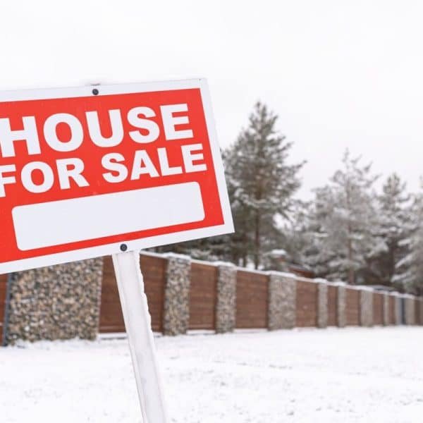 Un panneau « Maison à vendre » se dresse devant une cour enneigée et une clôture en bois, avec des arbres et un ciel nuageux en arrière plan.