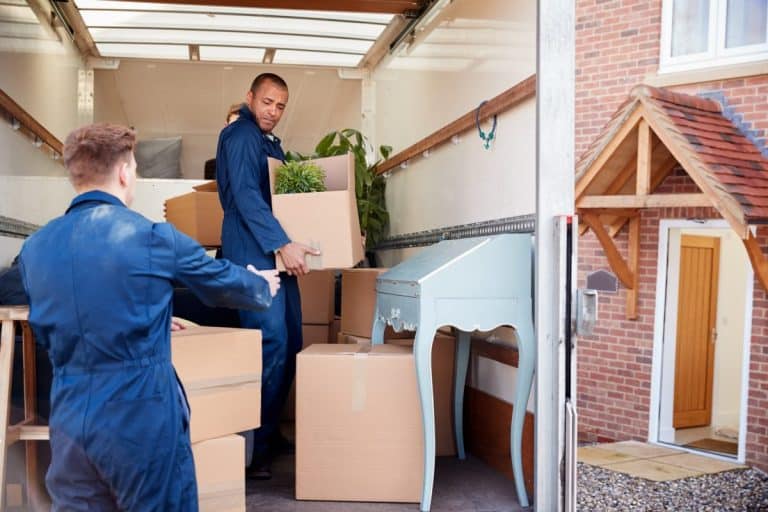 Deux déménageurs portant des uniformes bleus chargent des cartons et une petite table bleue dans un camion de déménagement garé devant une maison en briques avec une porte d'entrée en bois.