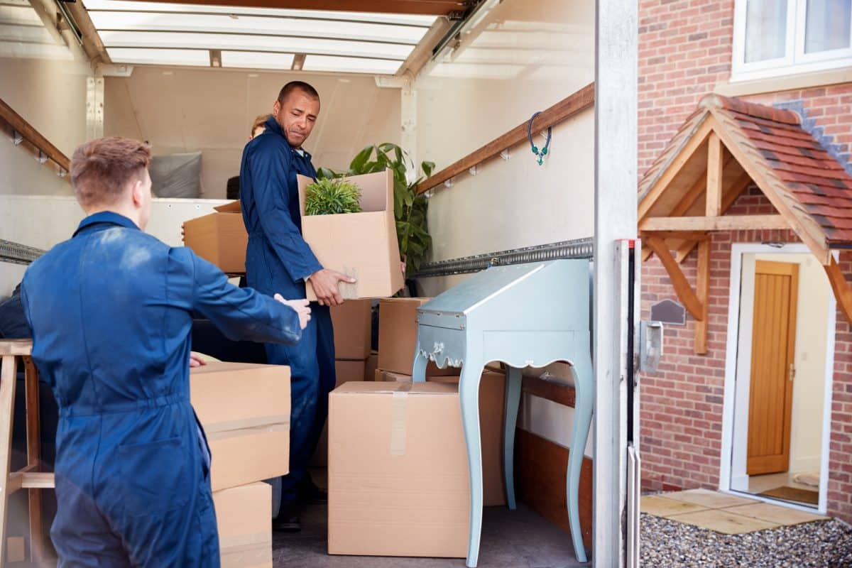 Deux déménageurs portant des uniformes bleus chargent des cartons et une petite table bleue dans un camion de déménagement garé devant une maison en briques avec une porte d'entrée en bois.