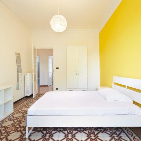 Chambre minimaliste avec un lit blanc, une armoire et une étagère sur un mur jaune vif. Un carrelage à motifs géométriques et un plafonnier rond rehaussent le style de la pièce.