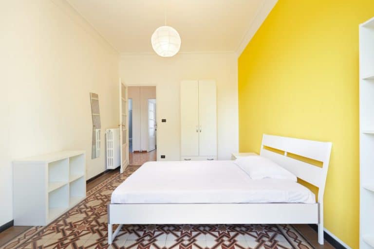 Chambre minimaliste avec un lit blanc, une armoire et une étagère sur un mur jaune vif. Un carrelage à motifs géométriques et un plafonnier rond rehaussent le style de la pièce.