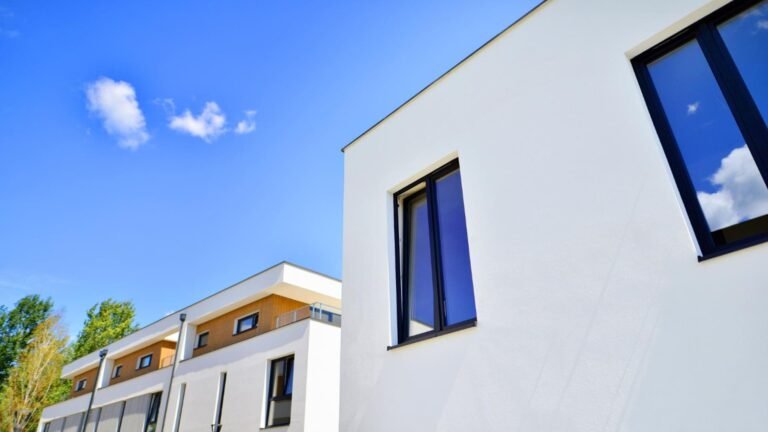 Bâtiments résidentiels modernes blancs avec de grandes fenêtres sous un ciel bleu clair, avec quelques nuages visibles en arrière-plan.