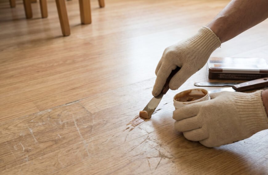 Gros plan sur des mains gantées appliquant une pâte de remplissage avec une spatule pour réparer des rayures sur un sol en lino clair.