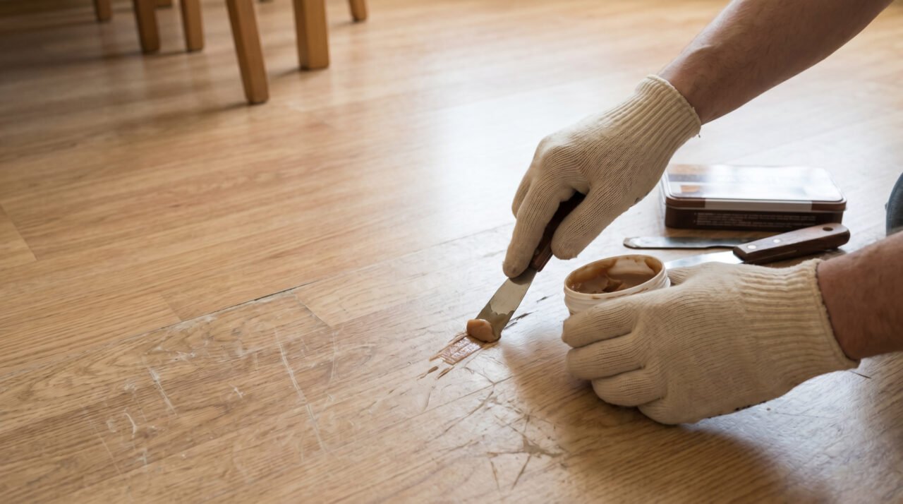 Gros plan sur des mains gantées appliquant une pâte de remplissage avec une spatule pour réparer des rayures sur un sol en lino clair.