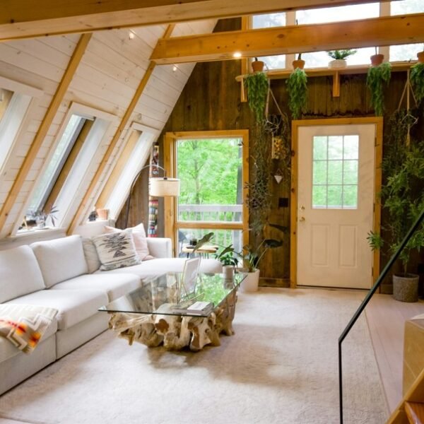 Salon en forme de A avec de grandes fenêtres, un canapé blanc, une table basse en verre, des plantes suspendues, des murs en bois et une guitare près d'un escalier.