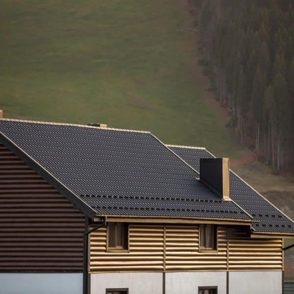 Une maison avec un toit sombre et un revêtement marron se trouve devant une colline herbeuse avec des arbres en arrière plan.