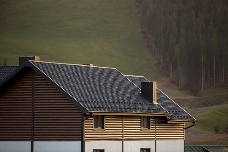 Une maison avec un toit sombre et un revêtement marron se trouve devant une colline herbeuse avec des arbres en arrière plan.