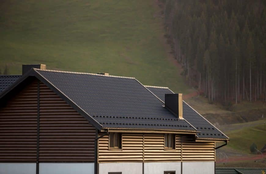 Une maison avec un toit sombre et un revêtement marron se trouve devant une colline herbeuse avec des arbres en arrière plan.