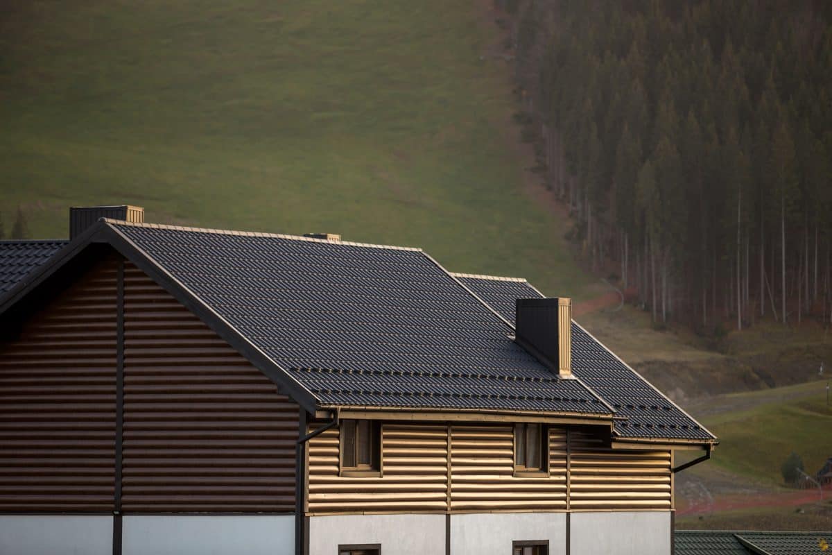 Une maison avec un toit sombre et un revêtement marron se trouve devant une colline herbeuse avec des arbres en arrière plan.