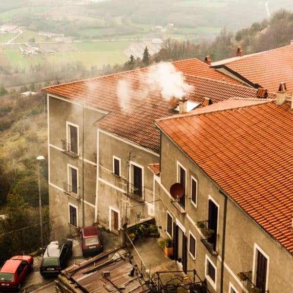 Trois voitures garées sur une colline escarpée à côté de maisons aux toits de tuiles rouges émettant de la fumée, au milieu d'un paysage rural brumeux avec des champs en arrière-plan.