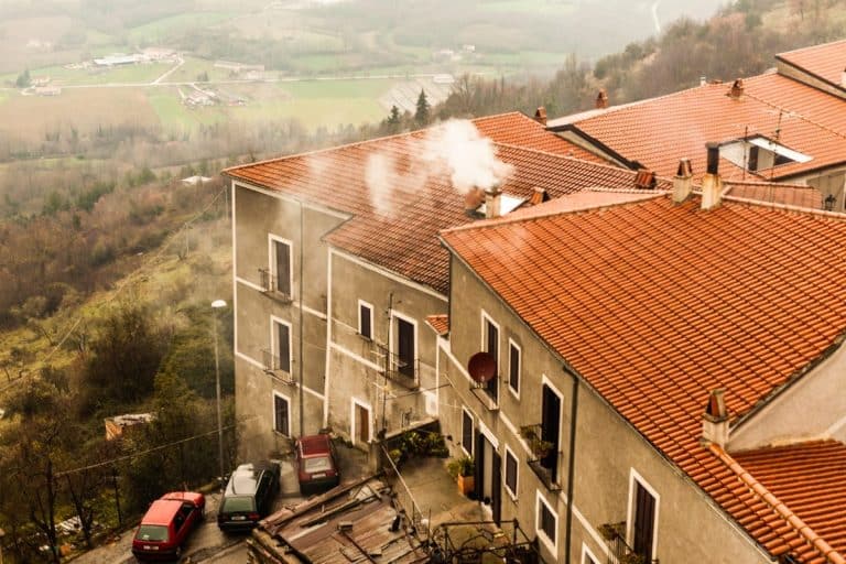Trois voitures garées sur une colline escarpée à côté de maisons aux toits de tuiles rouges émettant de la fumée, au milieu d'un paysage rural brumeux avec des champs en arrière-plan.
