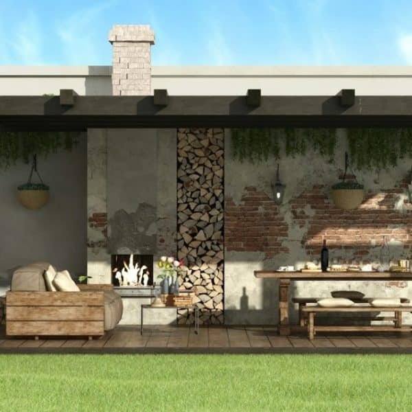 Terrasse extérieure avec pergola en bois, cheminée et coin repas. Comprend un canapé, une table, des plantes suspendues et du bois de chauffage empilé contre un mur de briques rustique, avec de la verdure entourant l'espace.