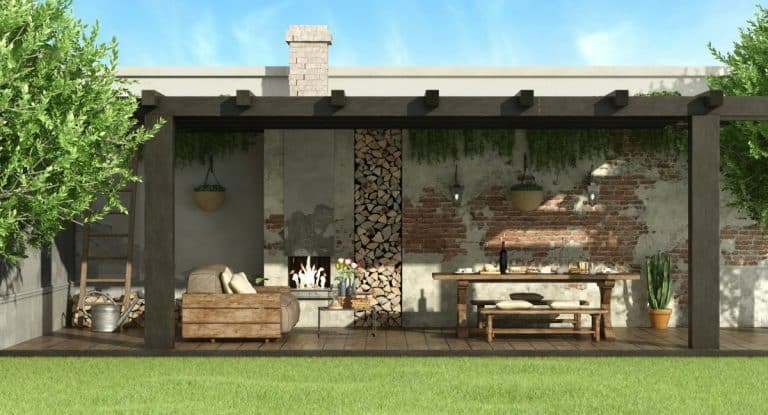 Terrasse extérieure avec pergola en bois, cheminée et coin repas. Comprend un canapé, une table, des plantes suspendues et du bois de chauffage empilé contre un mur de briques rustique, avec de la verdure entourant l'espace.
