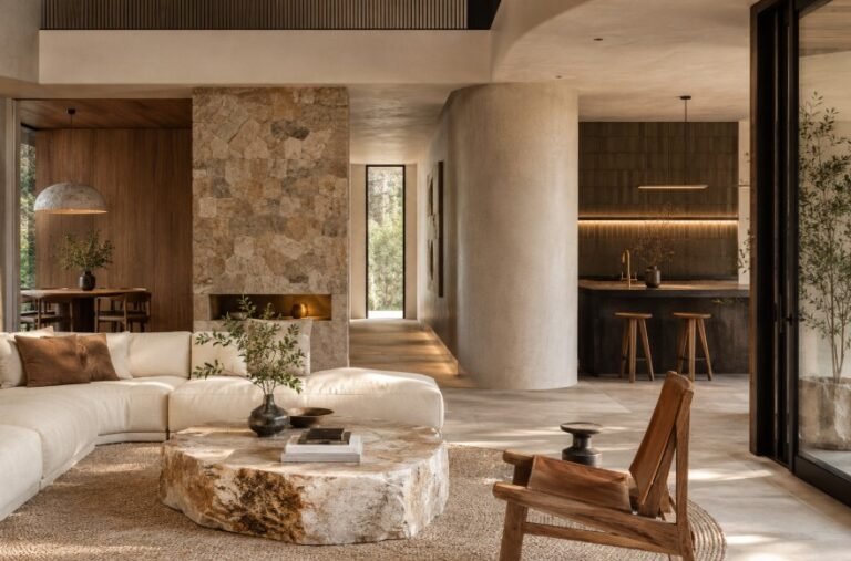 Salon moderne aux tons neutres, avec une table basse en pierre, un canapé modulable blanc, une chaise en bois, une cuisine ouverte et un coin repas, mettant en valeur des textures naturelles et de grandes fenêtres.