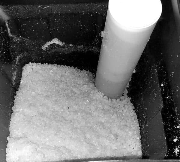 Récipient rempli de granulés de sel entourant un tube blanc vertical, faisant probablement partie d'un système d'adoucisseur d'eau.!