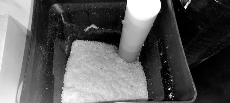 Récipient rempli de granulés de sel entourant un tube blanc vertical, faisant probablement partie d'un système d'adoucisseur d'eau.!
